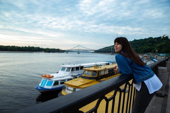 A young woman leaning on a railing overlooking docked boats and a suspension bridge during her Nile River Cruise journey