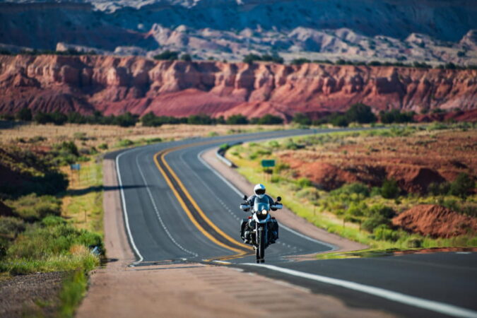 A spectacular wide-angle view of an empty highway curving through the heart of the Monument Valley desert, bathed in the golden light of a sunset. The iconic red rock buttes rise against the horizon, perfectly capturing the freedom and adventure expected during cross-country motorcycle tours.