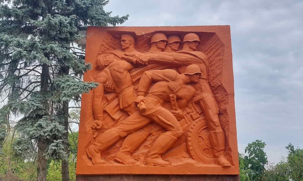 A terracotta-colored Soviet relief monument in Chi?in?u depicting dynamic figures of World War II soldiers in action, surrounded by evergreen trees under an overcast sky.