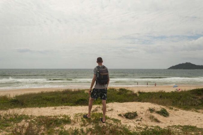 A man with a backpack stands on a grassy sand dune looking out at the ocean under a cloudy sky in "a Solo Island Traveler.jpg".