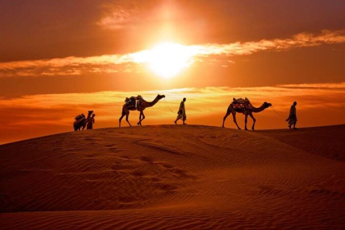 Silhouettes of people and camels walking along a ridge during a breathtaking desert sunset.